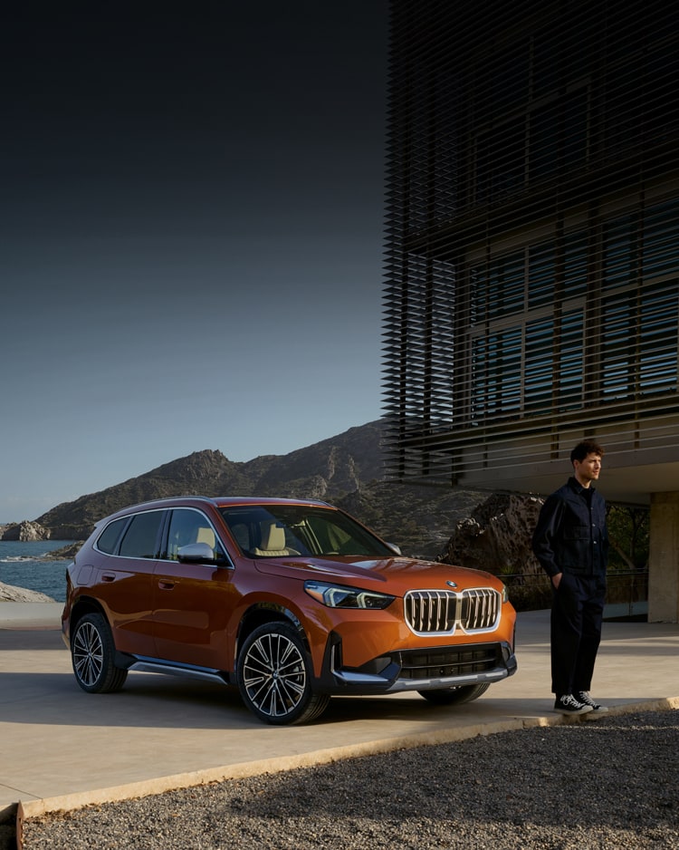 A casually-dressed man stands on a driveway in front of a parked BMW