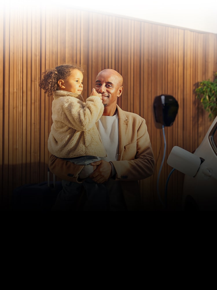 A smiling father holds his young daughter while charging his BMW at a home charger