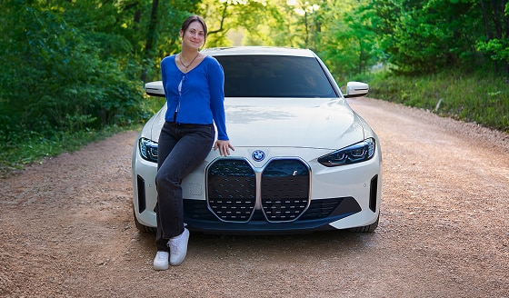 A smiling young woman leans on the front hood of a BMW i4 on a gravel road.