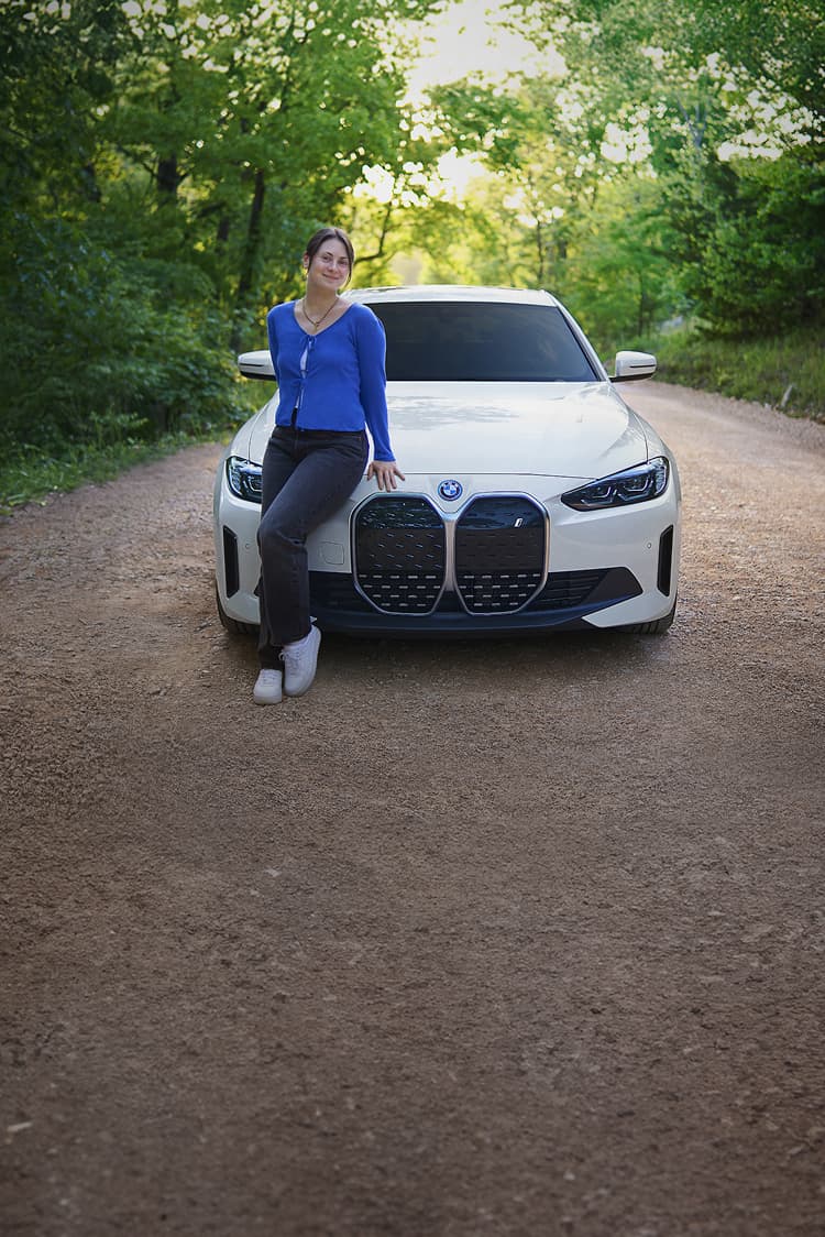 A smiling young woman leans on the front hood of a BMW i4 on a gravel road.